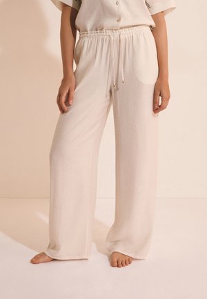 Personne portant un pantalon ample couleur crème avec cordon, debout pieds nus sur un fond beige clair.