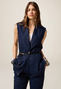 Navy sininen ilmanhiha blazer, jossa on muotoiltu istuvuus, yksittäinen nappikiinnitys ja musta vyö, joka korostaa vyötäröä. Silkkinen rakenne.