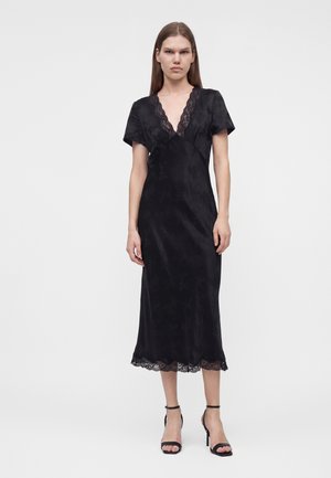 Femme aux longs cheveux raides portant une robe midi noire avec une bordure en dentelle au niveau de l'encolure et de l'ourlet, associée à des sandales noires à talons hauts.