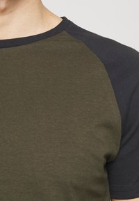 Olivově zelené bavlněné tričko s námořnicky modrými krátkými raglánovými rukávy. Hladká textura a kulatý výstřih. Jednoduchý design, bez vzorů nebo akcentů.