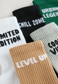 Cinq chaussettes côtelées en blanc, noir, vert, gris et marron, chacune avec des phrases motivationnelles en texte blanc ou noir, en gras.
