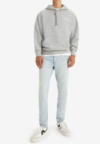 Mies, joka wears vaaleanharmaan Levi's hupparin, vaaleansiniset farkut, joissa on hieman kulutusta, valkoisen t-paidan ja valkoiset lenkkarit mustilla yksityiskohdilla.