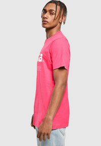 Camiseta de algodón de color rosa brillante con mangas cortas, que presenta un gran texto blanco en la parte frontal y un corte relajado. Costura visible en el dobladillo.