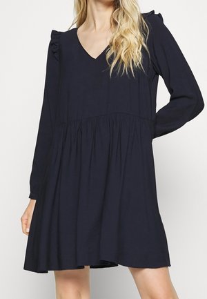 Kobieta w luźnej, długiej, czarnej sukience z długim rękawem, z dekoltem w serek i falbanami na ramionach, stojąca z jedną ręką na biodrze.