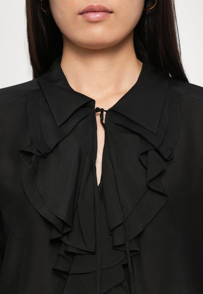 Woman wearing black blouse with large ruffled collar tied at the neck and gold hoop earrings, shown from chin to chest.