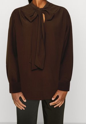 Kvinde iført en mørkebrun langærmet bluse med en stor bundet sløjfe ved halsudskæringen og sorte bukser, stående foran en ensfarvet baggrund.