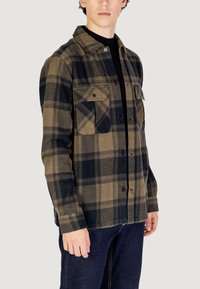Camicia di flanella a scacchi con grandi tasche, caratterizzata da sfumature di marrone e nero. Chiusura a bottone, colletto e vestibilità rilassata.