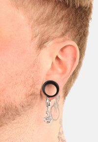 Svart silikon tunnelörhänge med en silverskorpion charm hängande från en ögla. Örat har ljusbrunt hår och synlig hudtextur.