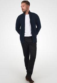 Marinblå, ribbet lynlåsjakke over en hvid T-shirt, parret med sorte teksturerede bukser og brune ankelstøvler. Model poserer med hænderne i lommen.