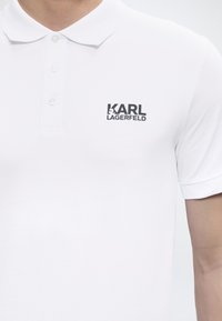 Λευκό πόλο με τρία κουμπιά, με μαύρο λογότυπο "KARL LAGERFELD" στο αριστερό στήθος, που φοράει ένα άτομο.
