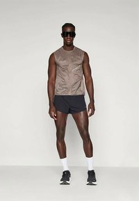 Άνδρας μοντέλο φορώντας αμάνικο τασάκι σε ταχύτερη απόχρωση, μαύρες αθλητικές βερμούδες, λευκές κάλτσες, μαύρα αθλητικά παπούτσια και μεγάλα μαύρα γυαλιά ηλίου, στέκεται μπροστά από απλό φόντο.