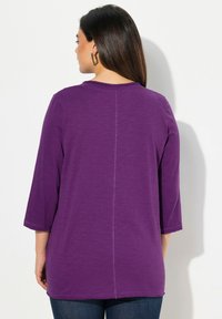 Lila, 3/4-Ärmel-Oberteil mit runder Halsauschnitt, hat eine mittige Rückennaht, besteht aus weichem, leichtem Stoff und wird mit dunkelblauen Jeans kombiniert.