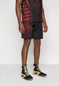 Μαύρα αθλητικά σορτς με κόκκινα γραφικά μοτίβα, συνδυασμένα με ταιριαστό τοπ. Μαύρα και μπεζ μποτάκια συμπληρώνουν την εμφάνιση.