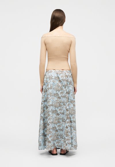 Γυναίκα με μακριά καστανά μαλλιά που φοράει μπεζ ριμπ πλεκτό τοπ χωρίς ώμους με κουμπιά και φούστα μάξι ανοιχτού μπλε με λουλουδάτο σχέδιο, που στέκεται μπροστά σε λευκό φόντο.