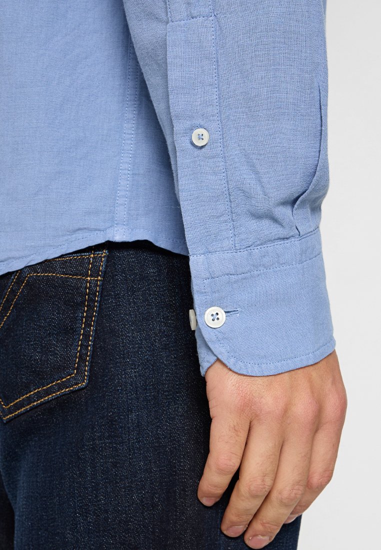 Camisa de manga larga azul claro con acabado texturizado, que muestra un puño abotonado y costuras detalladas, combinada con jeans de mezclilla oscuros.