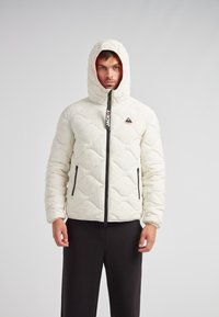 Puffjacka i off-white, med huva, quiltat mönster, svart dragkedja och fickor med dragkedja på sidorna. Logotyp patch på bröstet.