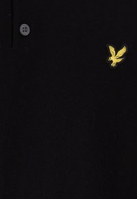 Svart stickad pikétröja med en gul broderad örnlogotyp på bröstet och en grå knapp vid kragen.