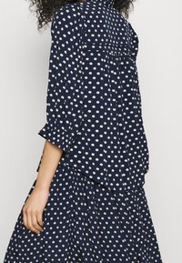 Marineblå blus med prickar, trekvartsärmar, lös passform och avskuren nederkant, tillverkad av lättviktigt tyg med en subtil textur.