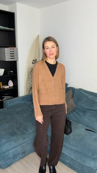 Γυναίκα που στέκεται σε εσωτερικό χώρο φορώντας καφέ ζακέτα, σκουρόχρωμο παντελόνι με σχέδια, μαύρα παπούτσια, κρατώντας μαύρη τσάντα κοντά σε μπλε καναπέ κοτλέ και σανίδα του σερφ.