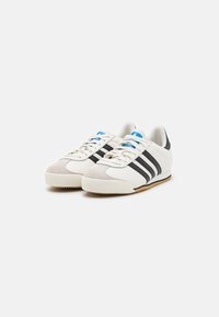 Chaussures de sport blanches et grises avec des rayures noires et des accents bleus. Elles sont fabriquées en matériaux en daim et synthétiques, avec une semelle en gomme texturée.