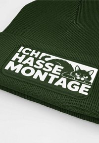 Green knit beanie featuring a white patch with the text "ICH HASSE MONTAGE" and a graphic of an annoyed cat. Text and graphics are bold and clear.