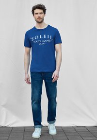 Sininen puuvillapaita, jossa on valkoista tekstiä, yhdistettynä sinisiin farkkuihin ja valkoisiin tennareihin. Rentoa istuvuutta ja lyhyet hihat.