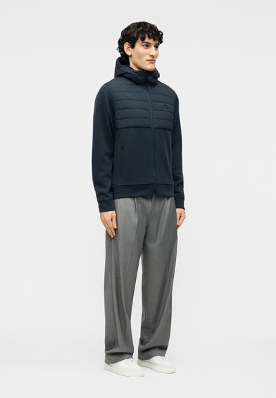 Νέος άντρας που στέκεται, φοράει μια σκούρα μπλε καπιτονέ ζακέτα με φερμουάρ, φαρδιά γκρι παντελόνια και λευκά αθλητικά παπούτσια μπροστά σε ένα απλό φωτεινό φόντο.