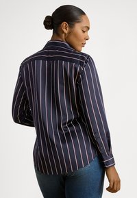 Žena s vlasmi zviazanými do drdolu, nosiaca kruhové náušnice a tmavomodrú košeľu s bielymi vertikálnymi pruhmi na dlhé rukávy, v kombinácii s modrými džínsami, otočená nabok.