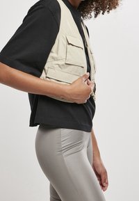 Beżowa kamizelka cargo z wieloma kieszeniami, zapinana na zamek, noszona na czarnej koszulce, zestawiona z błyszczącymi szarymi legginsami. Gładka tekstura, casualowy design.