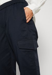Personne portant un pantalon cargo bleu marine avec une poche à rabat, une main dans la poche latérale et une chemise blanche à manches longues.