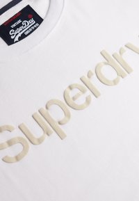 Hvid t-shirt med et cremefarvet, tekstureret "Superdry" logo, der er tydeligt vist på forsiden. Rund hals og blødt bomuldsstof.
