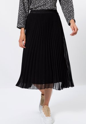 Schwarzer plissierter Midirock mit transparentem Stoff und elastischem Bund, kombiniert mit hellfarbenen Sneakern für einen lässigen Look.