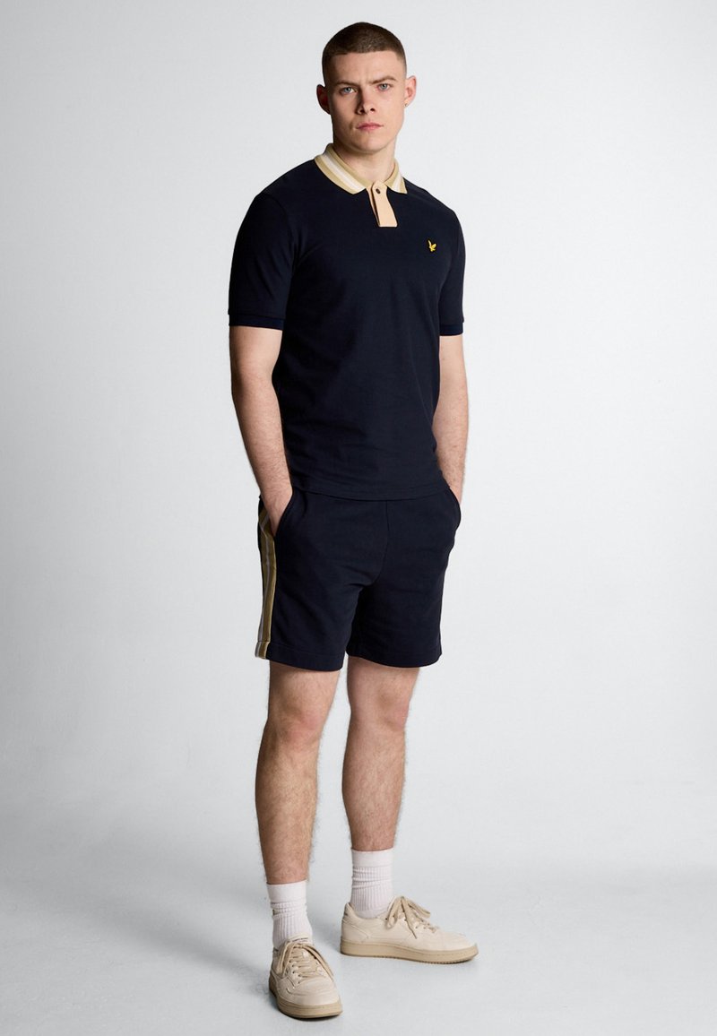 Polo shirt blu navy con colletto beige, dotato di un logo giallo. Abbinato a pantaloni corti blu navy con strisce laterali beige e sneakers beige.