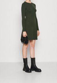Robe vert olive à manches longues avec un corsage ajusté et une jupe évasée, associée à des bottines noires épaisses et un petit sac à main noir.