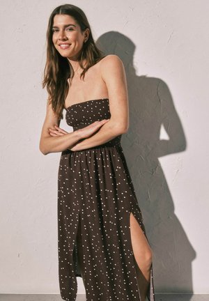 Junge Frau mit langen Haaren lächelt, trägt ein braunes trägerloses Kleid mit weißen Punkten und Seitenschlitzen, steht vor einer strukturierten Wand.