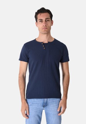 Uomo con capelli corti e scuri che indossa una camicia blu navy a maniche corte con tre bottoni e jeans azzurri, in piedi davanti a uno sfondo semplice.