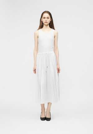 Junge Frau steht nach vorne blickend, trägt ein ärmelloses weißes Kleid mit plissiertem Rock und schwarze High-Heels vor weißem Hintergrund.