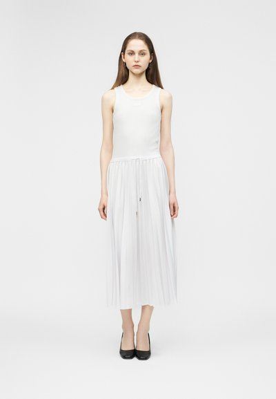 Jeune femme debout de face portant une robe blanche sans manches avec une jupe plissée et des chaussures à talons noirs sur un fond blanc.