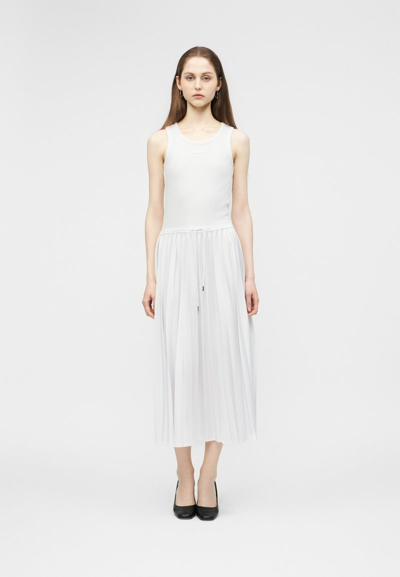 Νεαρή γυναίκα στέκεται προς τα εμπρός φορώντας ένα λευκό αμάνικο φόρεμα με πλισέ φούστα και μαύρα ψηλοτάκουνα παπούτσια σε λευκό φόντο.