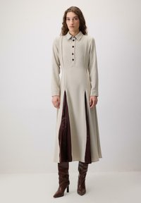 Lang, beige kjole med krave, knaplukning foran og to dybe burgundy slids i siderne, parret med brune hælsko. Glat tekstur.