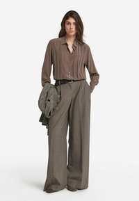 Långärmad, knappad blus i brunt med vertikala veck, kombinerad med vida grå byxor och en texturerad grå jacka hängande över en arm.