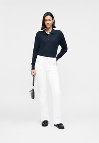 Μπλε πόλο με υφή και μακριά μανίκια, με λευκό γιακά, συνδυασμένο με λευκό παντελόνι με διακοσμητικά κουμπιά και μαύρα παπούτσια, κρατώντας μια μικρή τσάντα με σχέδιο.