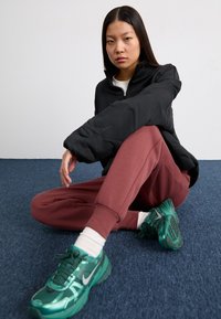 Casaco acolchoado preto com fecho, calças de moletom bordô, ténis verdes da Nike, meias brancas. Sentado contra um fundo de carpete azul.