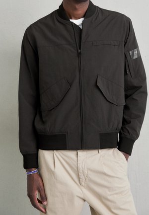 Mand iført sort , lynlåslukket bomberjakke med klaplommer og beige bukser, stående med den ene hånd i lommen mod en ensfarvet væg.