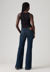 Femme aux cheveux tressés portant un top noir sans manches et un jean évasé bleu foncé, debout avec les mains sur les hanches, de dos sur un fond uni.