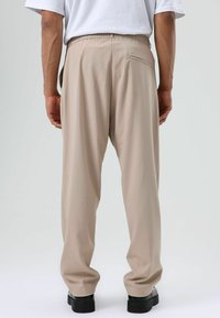 Beige, loszittende broek met een elastische tailleband, voorzien van een achterzak en een gladde textuur, gecombineerd met zwarte schoenen.