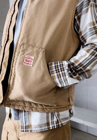 Inimene kanda beeži töövesti ja ruudulise särgiga, käsi vestitaskus, näidates Levi Strauss & Co. silti taskus.