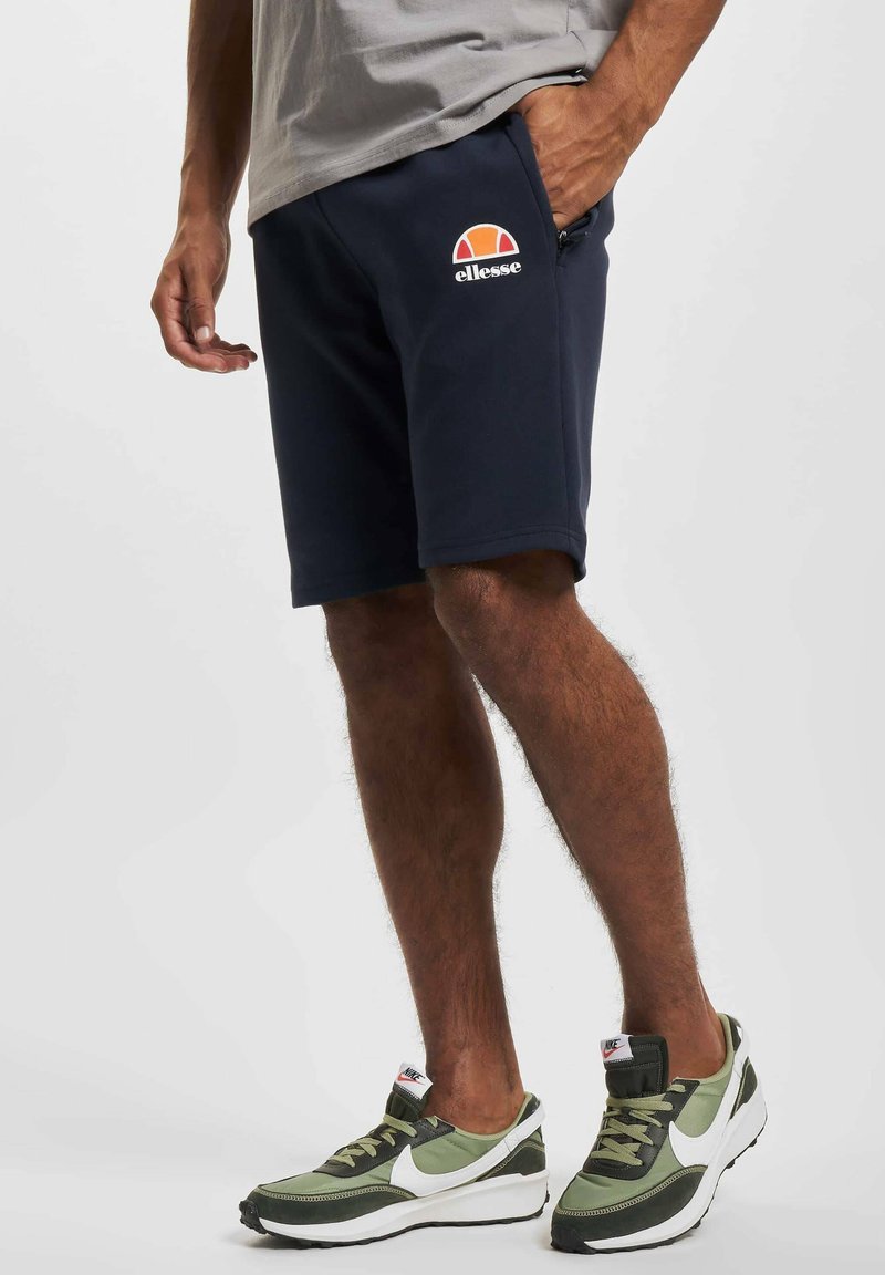 Pantalones cortos azul marino de tela suave, con un logo de Ellesse en la parte delantera y bolsillos laterales. Combinados con zapatillas deportivas verdes y blancas.