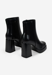 Botines negros de tobillo hechos de cuero brillante con un alto tacón bloque y cierre de cremallera. Presentan una superficie lisa y sólida con punta redondeada.
