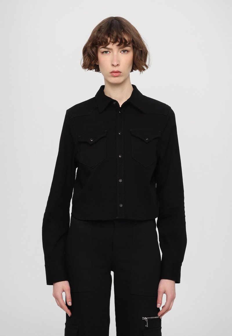Camicetta nera corta con bottini sul davanti, due tasche sul petto, colletto all'uncinetto e maniche lunghe. Tessuto liscio e colore uniforme.